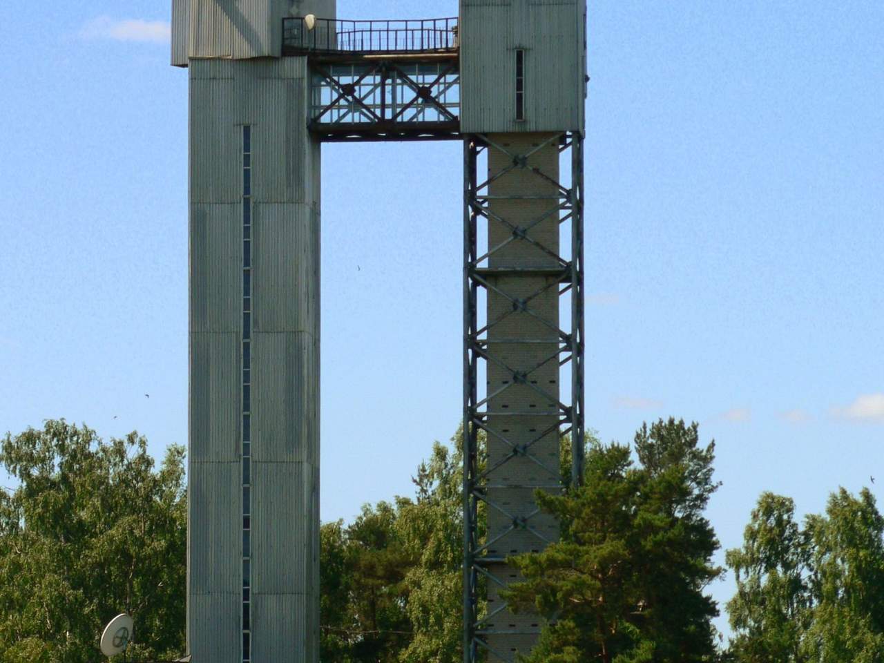 Aukštas betoninis bokštas su metaline konstrukcija viršuje po giedru dangumi tarp medžių