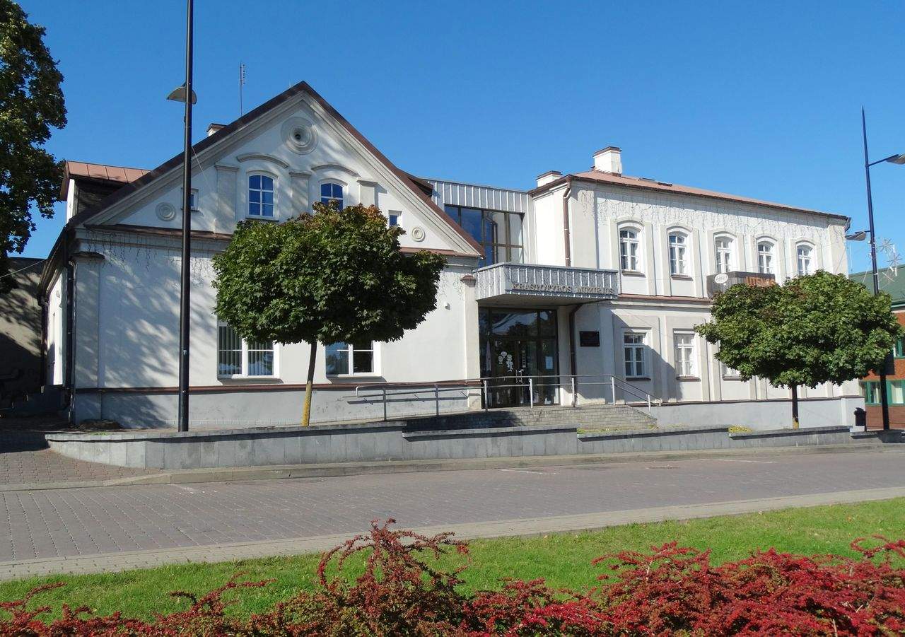 Šviesus Utenos kraštotyros muziejaus pastatas su dviem aukštais ir vešliais medžiais priekyje